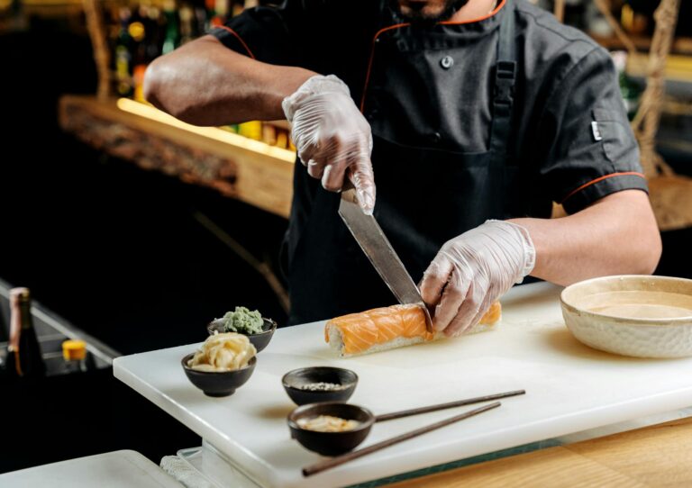 Chef wearing gloves slices sushi rolls on a chopping board in a restaurant kitchen.